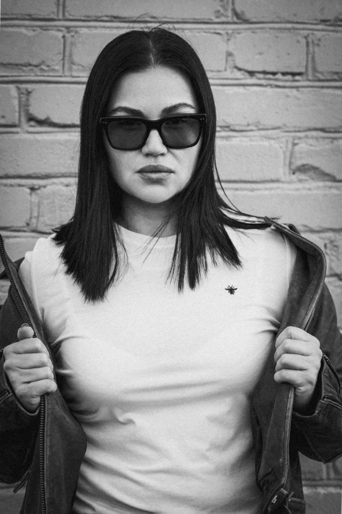 Чёрно-белый портрет у кирпичной стены: очки, белая футболка, тёмная куртка. Ольга Грачёва